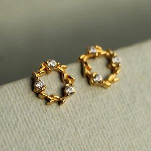 Side angle of gold wreath circle stud earrings with intricate leaf motif and embedded cubic zirconia stones resting on a pale display board.