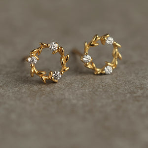 Top-down view of dainty gold-plated wreath earrings with laurel leaf shapes and shimmering cubic zirconia detailing on a grey stone background.