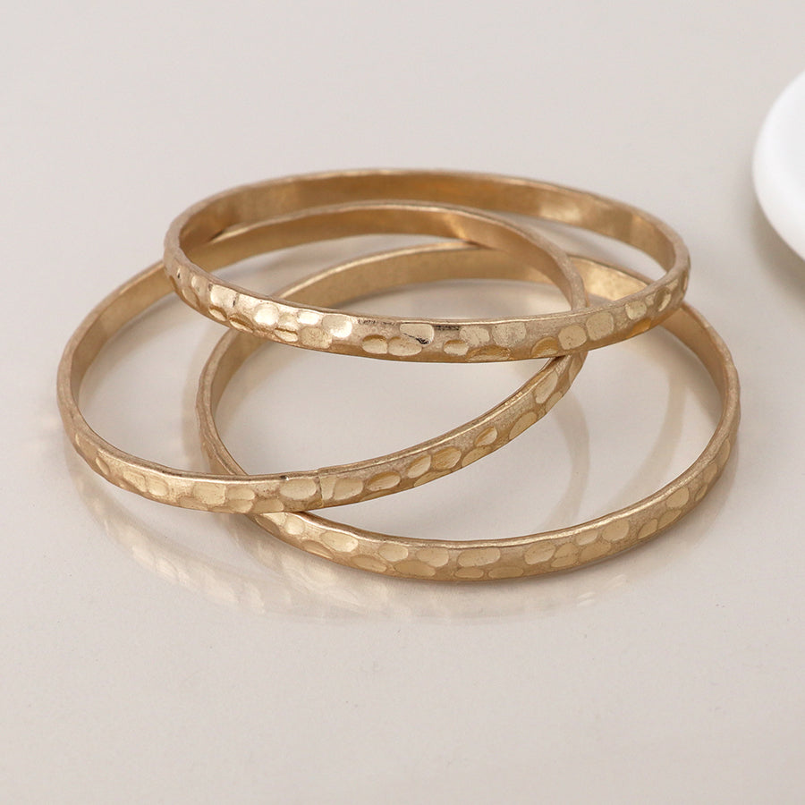 Trio of gold hammered bangles laid flat on neutral background.
