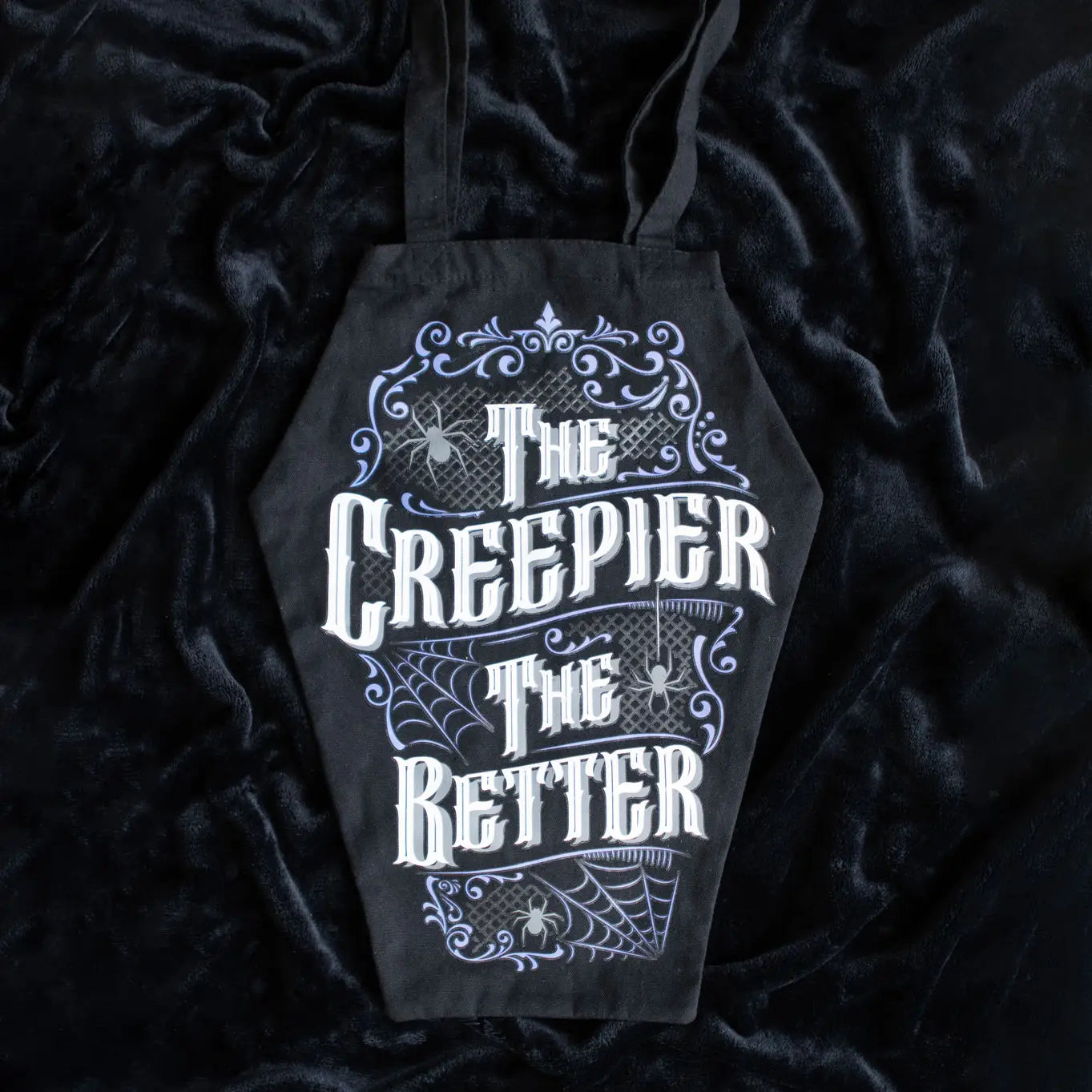 Gothic coffin-shaped black tote bag with white “The Creepier The Better” text and purple spiderweb design, displayed on black velvet fabric.