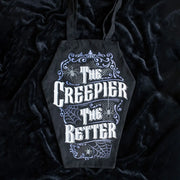 Gothic coffin-shaped black tote bag with white “The Creepier The Better” text and purple spiderweb design, displayed on black velvet fabric.