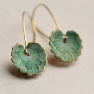 A pair of handmade green clover leaf earrings with gold-tone detailing, displayed on a neutral surface. The brass charms are painted and varnished for depth and detail.