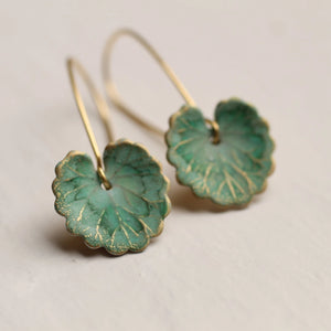 Close-up of the green clover leaf earrings, showing the intricate veining and gold accents on the brass charms. The earrings hang from delicate brass ear wires.