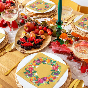 Autumn-themed table setting featuring a green glass candlestick holder with a dark green candle, surrounded by fruits and gold-accented napkins.