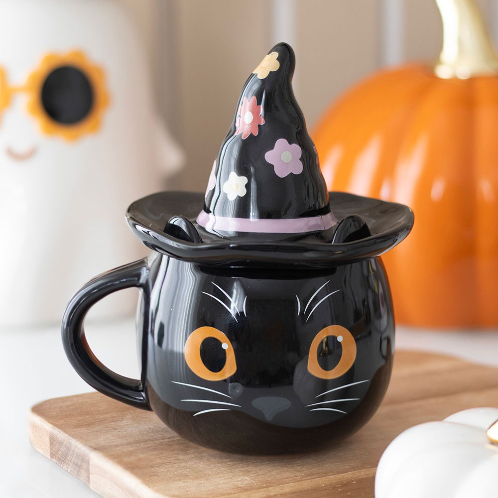 Front view of black cat mug with floral witch hat lid on a wooden tray beside autumn decorations.