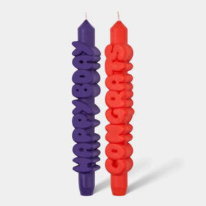 Front-facing view of a sculptural candle set with the words “Happy Bday” in indigo and “Congrats” in bright orange, designed in chunky 3D typography.