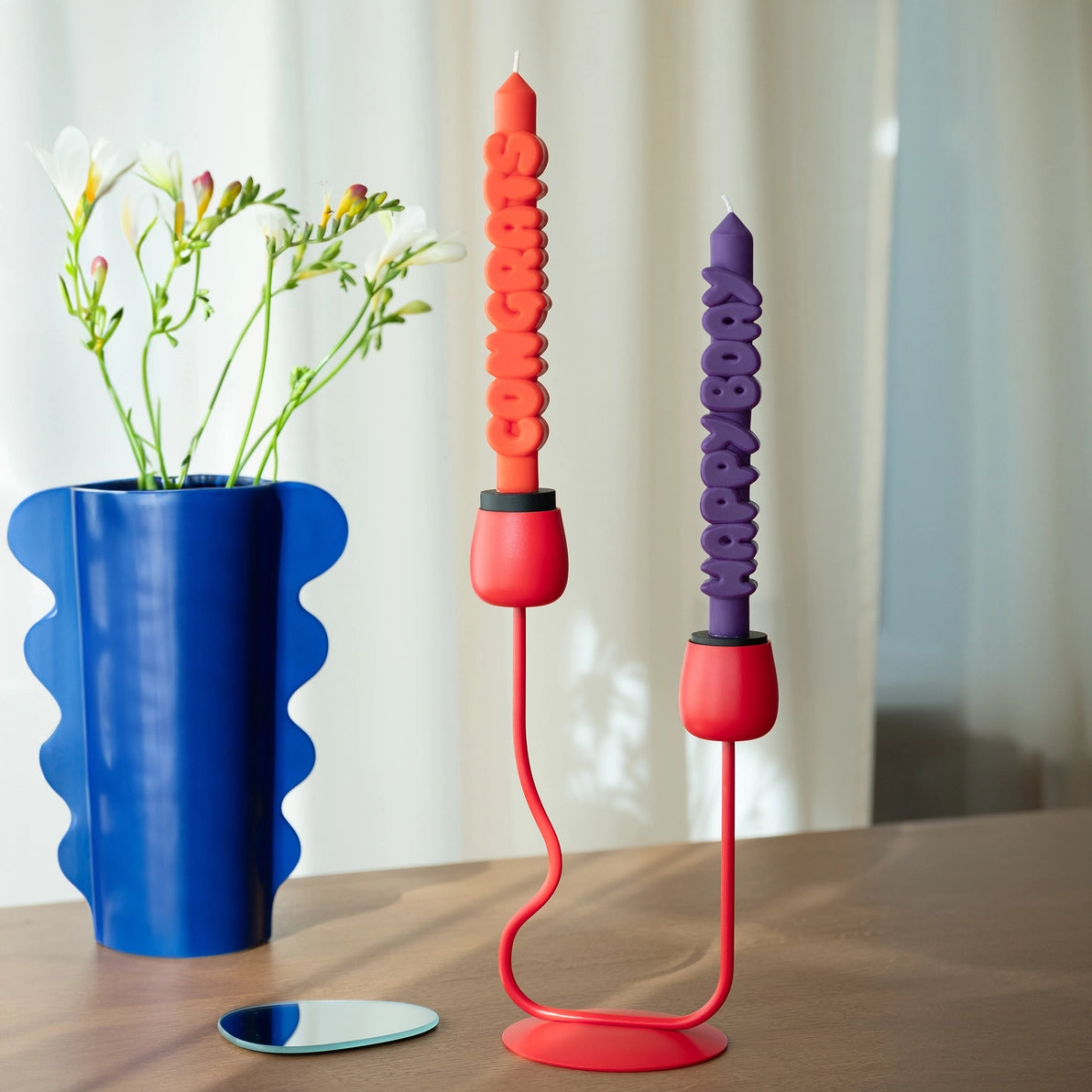 Indigo “Happy Bday” and orange “Congrats” taper candles displayed in a red modern double candle holder on a wooden table next to a blue wavy-edged vase with white flowers.