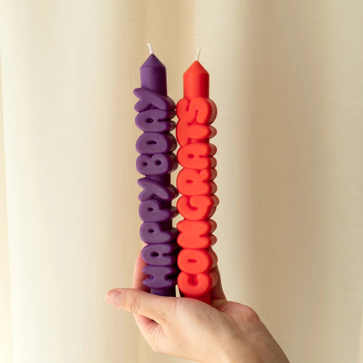 Hand holding two vibrant typographic taper candles with bold 3D lettering—one indigo with “Happy Bday” and one orange with “Congrats”—against a neutral background.