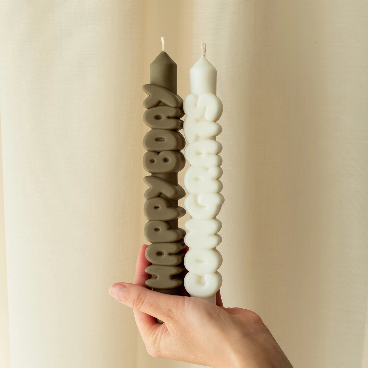 Hand holding a pair of novelty taper candles, featuring “Happy Bday” in olive green and “Congrats” in white, against a softly lit cream curtain backdrop.