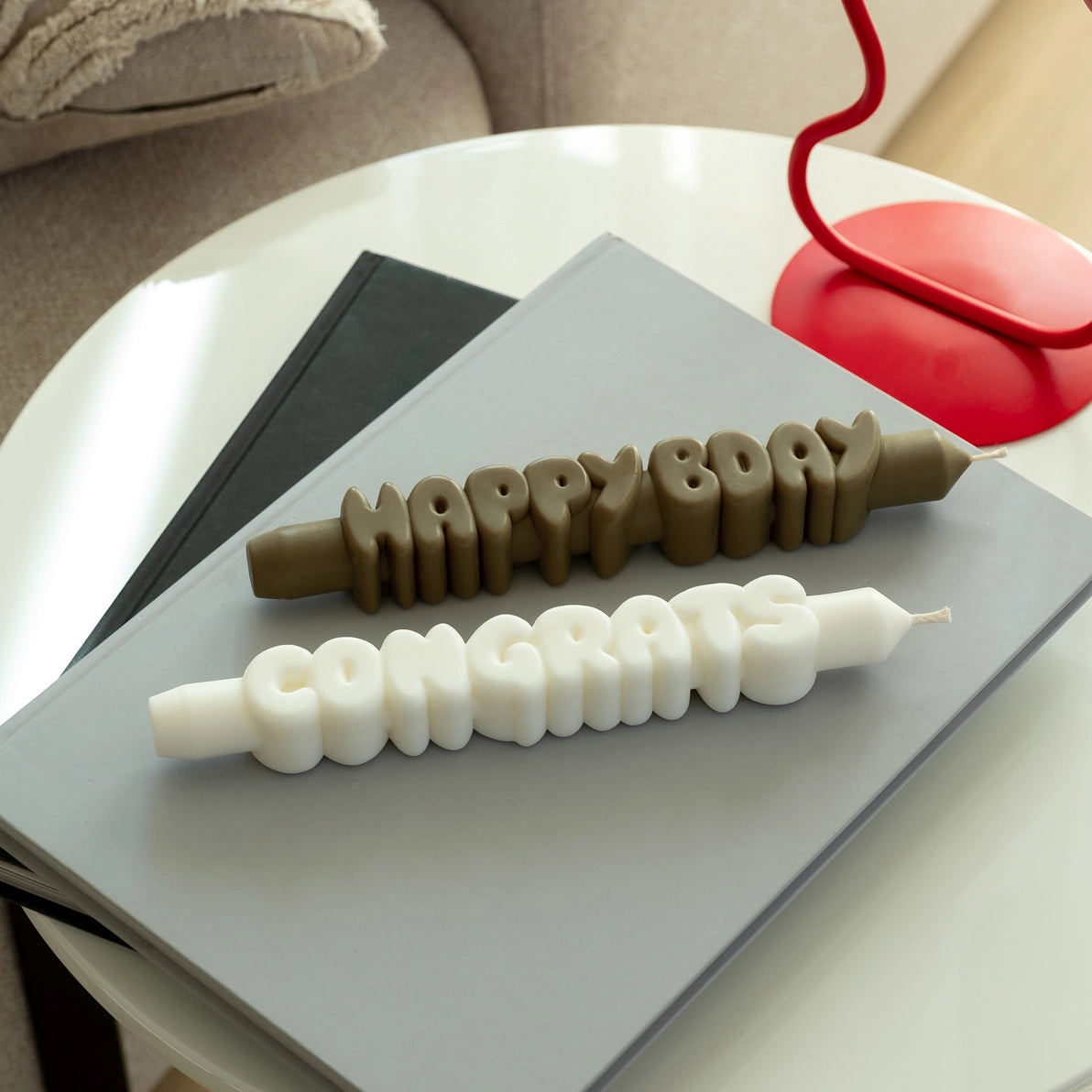 Overhead view of olive green “Happy Bday” and white “Congrats” candles lying side by side on a coffee table with stacked books in a modern home setting.