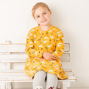 Young girl wearing Harry Potter™ Hedwig Dress — mustard yellow with all-over magical print, sitting on white bench.