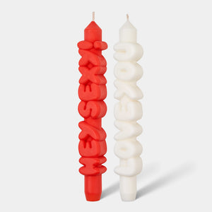 Front-facing view of two sculptural taper candles, one red reading “Hey Sexy!” and one white reading “Love You”, standing upright on a white background.