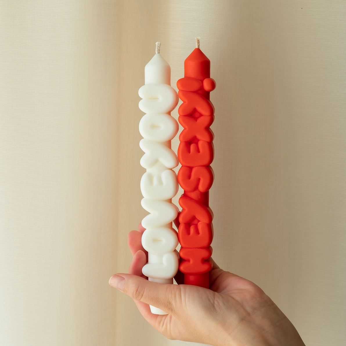 A hand holding two novelty taper candles—red “Hey Sexy!” and white “Love You”—against a neutral fabric backdrop, showcasing their bold 3D lettering.