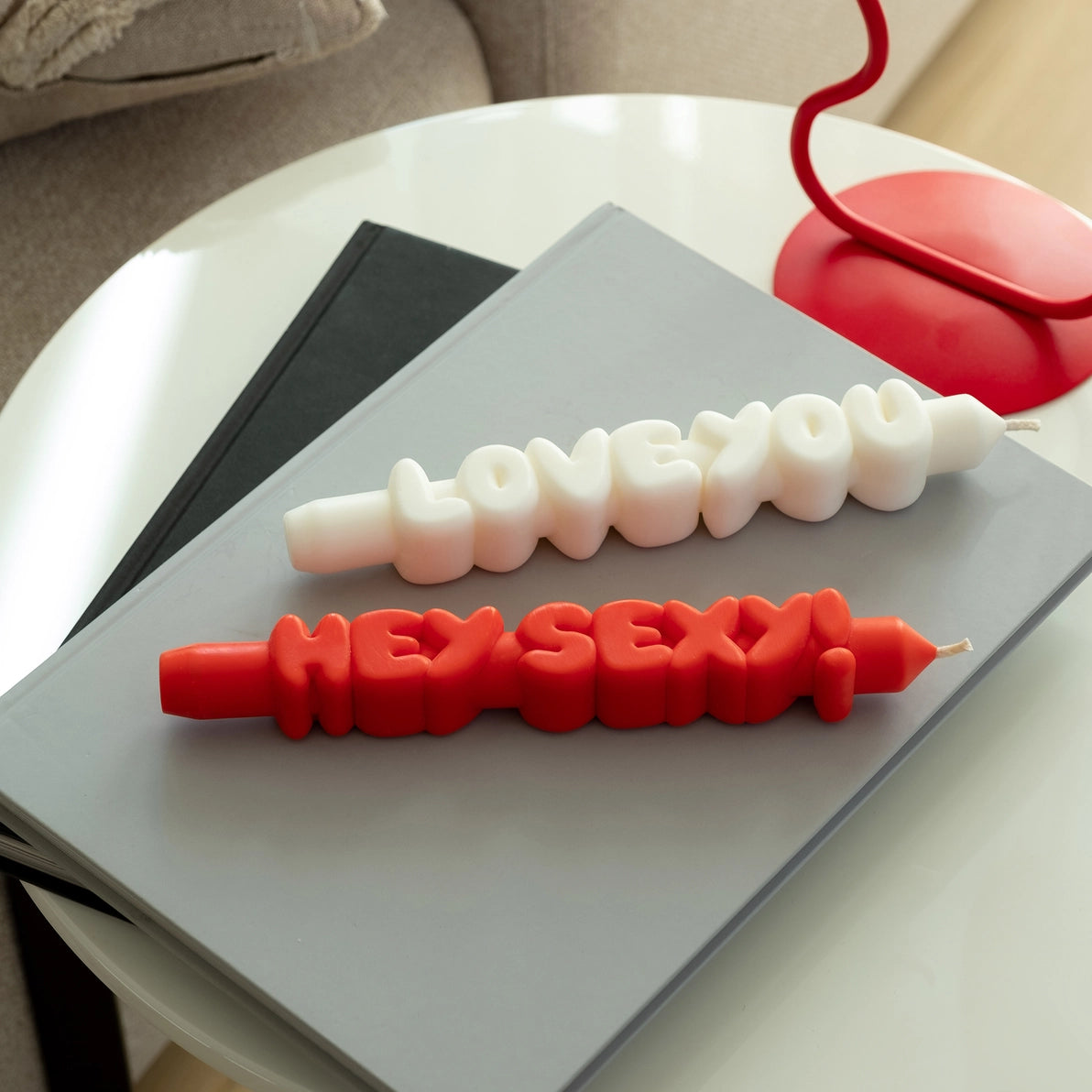 Overhead view of sculpted “Hey Sexy!” red candle and “Love You” white candle lying flat on a table next to books and a red lamp, styled in a cosy home interior.
