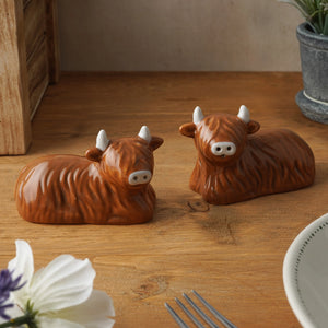 Ceramic Highland Cow salt and pepper shakers on wooden table with tableware and plants – rustic kitchen styling