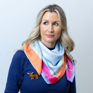 A woman with long blonde hair wears a vibrant coral and blue Hairy Coo scarf draped around her neck, paired with a navy blue top. A small handcrafted Highland cow brooch is pinned to her chest.