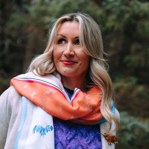 A woman with long blonde hair smiles while wearing a coral and blue Hairy Coo scarf draped over a warm purple knit sweater. The scarf features delicate cow illustrations, and a handcrafted Highland cow brooch is attached to her coat.