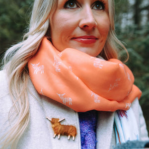 A close-up of a woman wearing a coral Hairy Coo scarf wrapped around her neck, with white cow illustrations woven into the design. A small handcrafted Highland cow brooch is pinned to her coat, adding a charming touch.