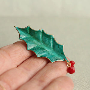 Verdigris enamel holly leaf brooch with red bead berries shown resting on an open palm for scale.