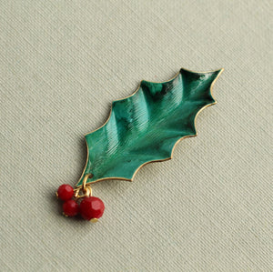 Angled view of vintage-style holly brooch featuring detailed green leaf texture, gold edging, and three red berries.