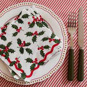 Christmas place setting with Holly White napkin folded on a white and red plate, decorated with holly leaves, red berries, and red wavy trim, styled on a candy stripe tablecloth.