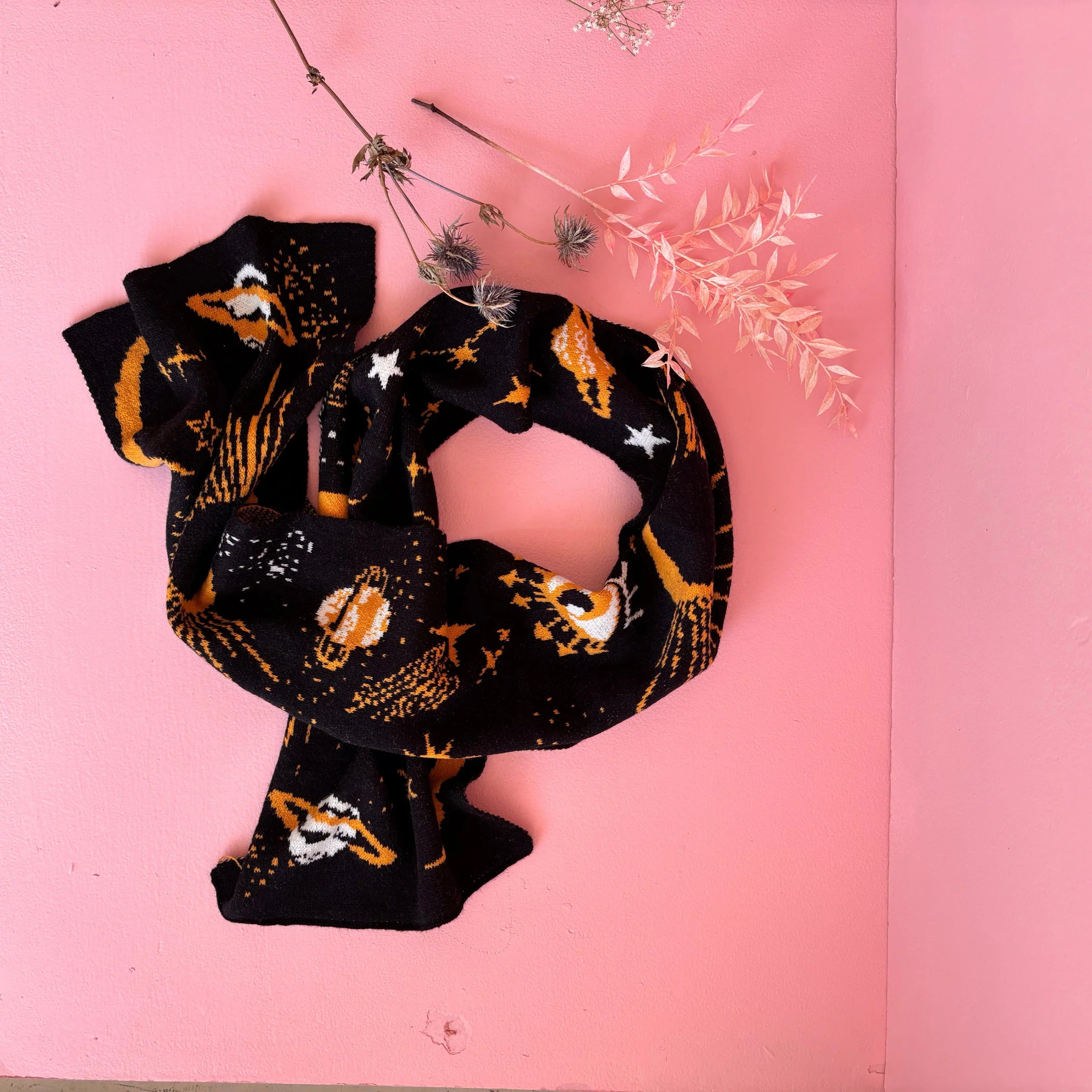 After Dark scarf displayed in a loop on a pink surface, showing the celestial-themed knit design with stars, planets, and suns.