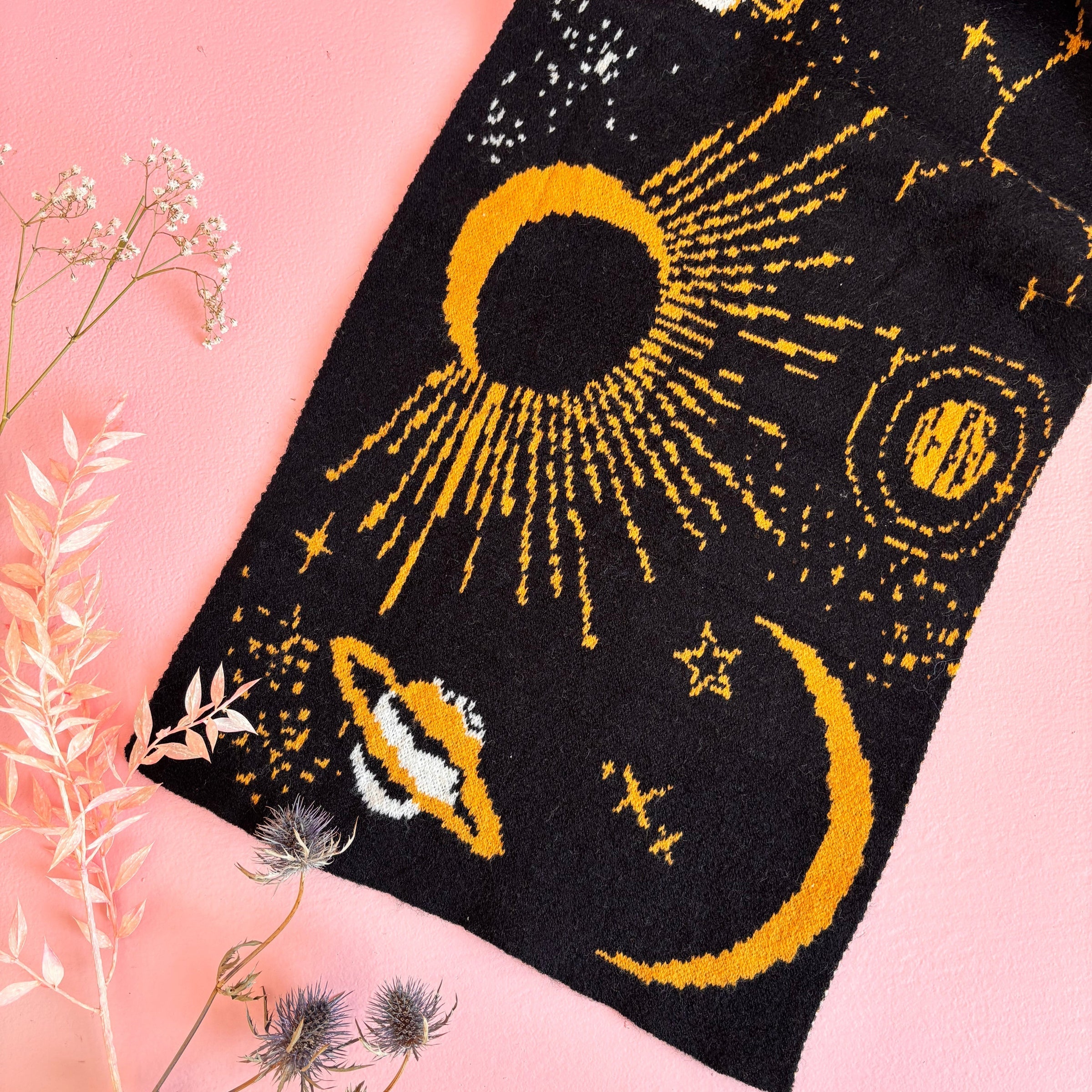 Close-up flat lay of eclipse and crescent moon design woven into black scarf with golden threads, surrounded by botanical decor.