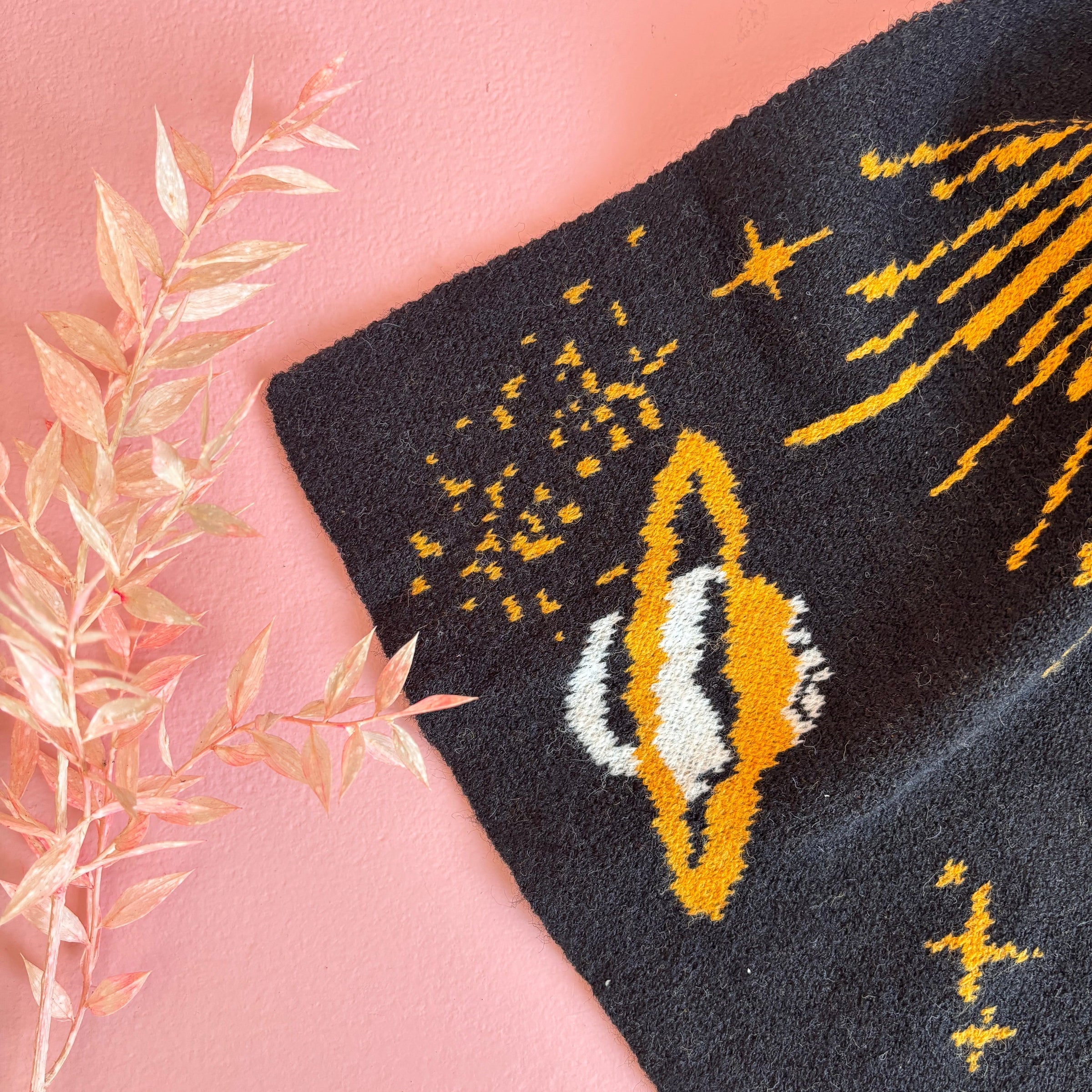 Flat lay close-up of golden Saturn-style planet detail on black knit scarf with pink background and dried floral styling.