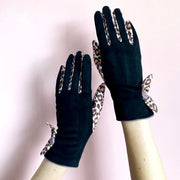 Black winter gloves with leopard print bow detail on wrists, shown against pale pink background.