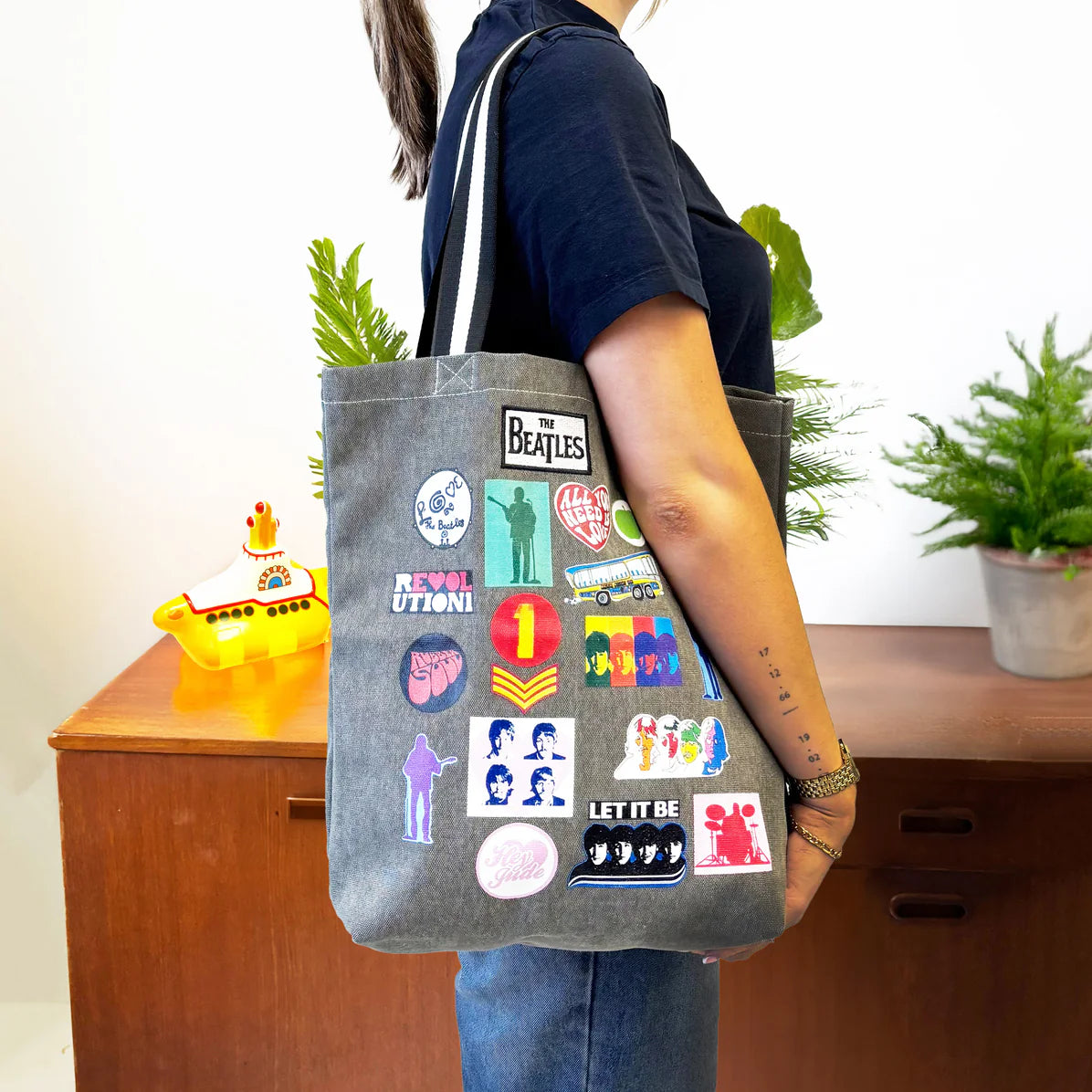 Side view of Beatles canvas tote bag worn on shoulder, showing patch prints and striped straps against a retro wooden backdrop.