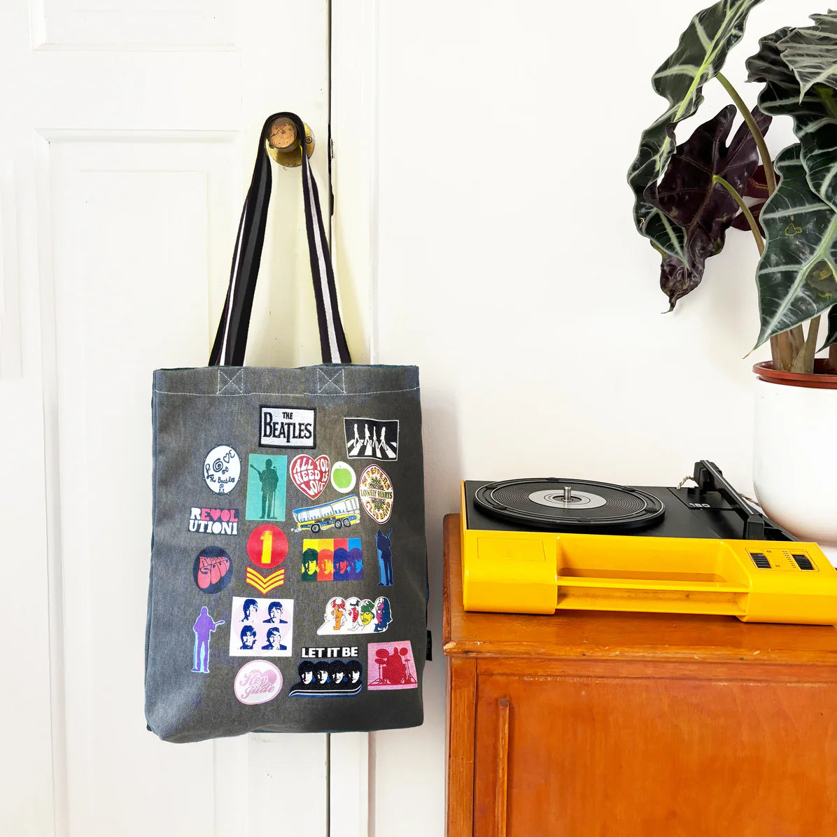 The Beatles tote bag hanging from a door with a record player nearby, showcasing colourful music-themed patches on grey canvas.