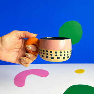 Hand holding Bloc Cup with terracotta handle and pink ceramic body against colourful abstract background.