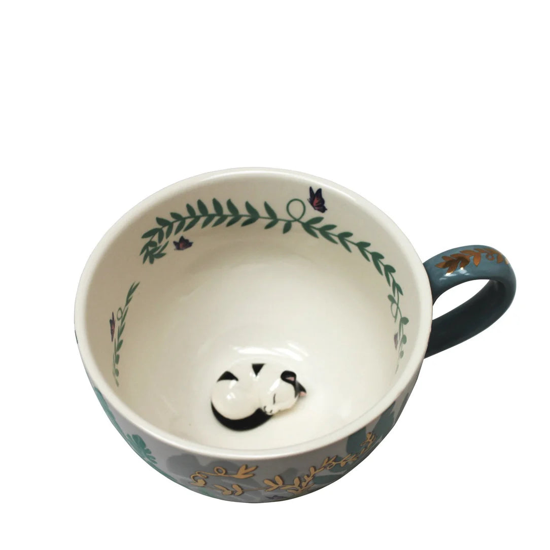 Angled view into porcelain cup showing curled up black-and-white cat at the bottom.