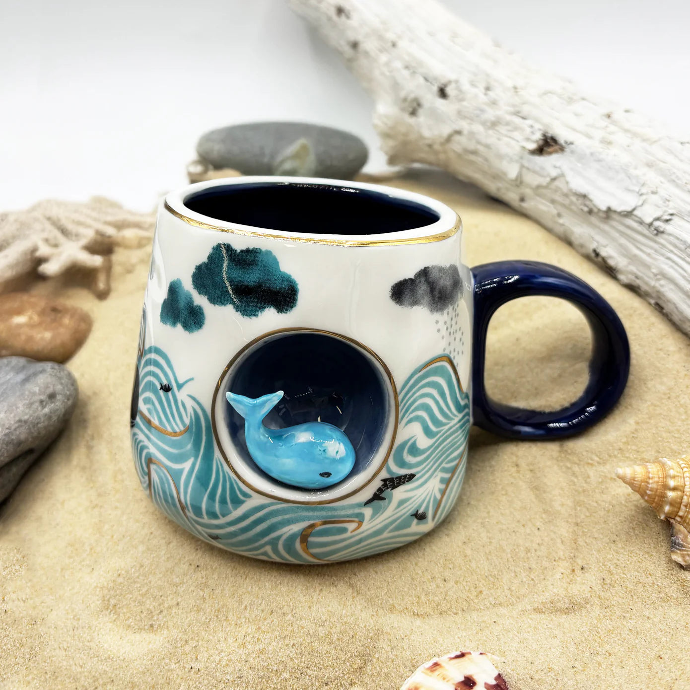 By The Sea ceramic mug with 3D whale and hand-painted ocean wave and storm design, styled on sand with shells.