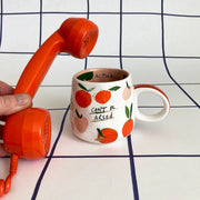 Close-up of ceramic “Can’t Be Arsed” mug with peach pattern, shown next to a bright orange retro-style telephone handset.
