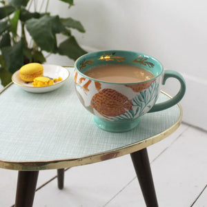 Large turquoise ceramic mug with gold coral detail and illustrated fish, filled with tea and placed on a retro side table next to a yellow macaron.