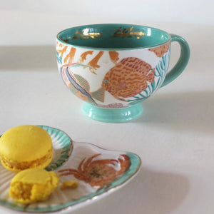 Coral-themed porcelain mug with sea life illustrations and gold rim, styled with yellow macarons on a crab-decorated seashell plate.