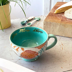 Interior view of the Coral Fish Mug showing turquoise glaze and gold coral design, placed on a rustic kitchen table with bread and cheese.