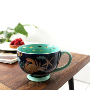 Porcelain Coral Lobster Cup on wooden table, featuring deep turquoise interior with goldfish motifs and a miniature ceramic lobster at the bottom.