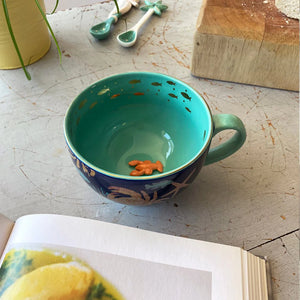 Coral Lobster Cup on a rustic surface with cookbook, highlighting the turquoise interior, golden fish design, and mini ceramic lobster.