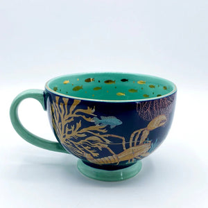 Side angle of Coral Lobster Cup featuring a golden lobster, seaweed, and fish against a dark blue porcelain background.