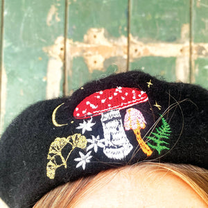 Close-up of embroidered mushroom design on black wool beret, featuring red and pink toadstools, golden leaves, green fern, and stars – Forage collection detail.