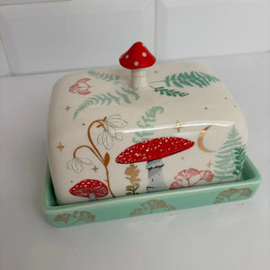 Side angle of the porcelain toadstool butter dish highlighting hand-painted ferns, white wildflowers, and gold moon detailing on a cream lid with green base.