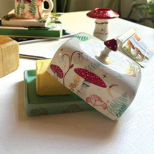Angled shot of the butter dish lid being removed, showing creamy ceramic surface with red toadstools, gold stars, and floral illustrations.
