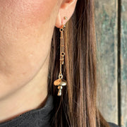 Close-up of a woman’s ear wearing a long gold drop earring with a mushroom charm and delicate chain, styled with straight brown hair.