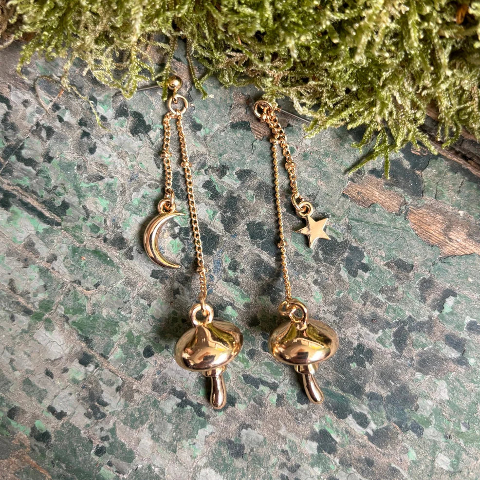 Gold mushroom drop earrings with crescent moon and star charms, displayed on a green rustic surface with moss decoration.