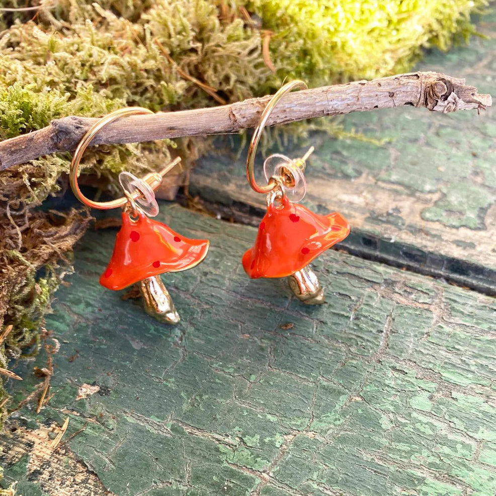 Close-up of Forage Red Mushroom Earrings hanging on a branch, featuring glossy red caps with subtle polka dots and gold-plated stems, styled against moss and rustic green wood.