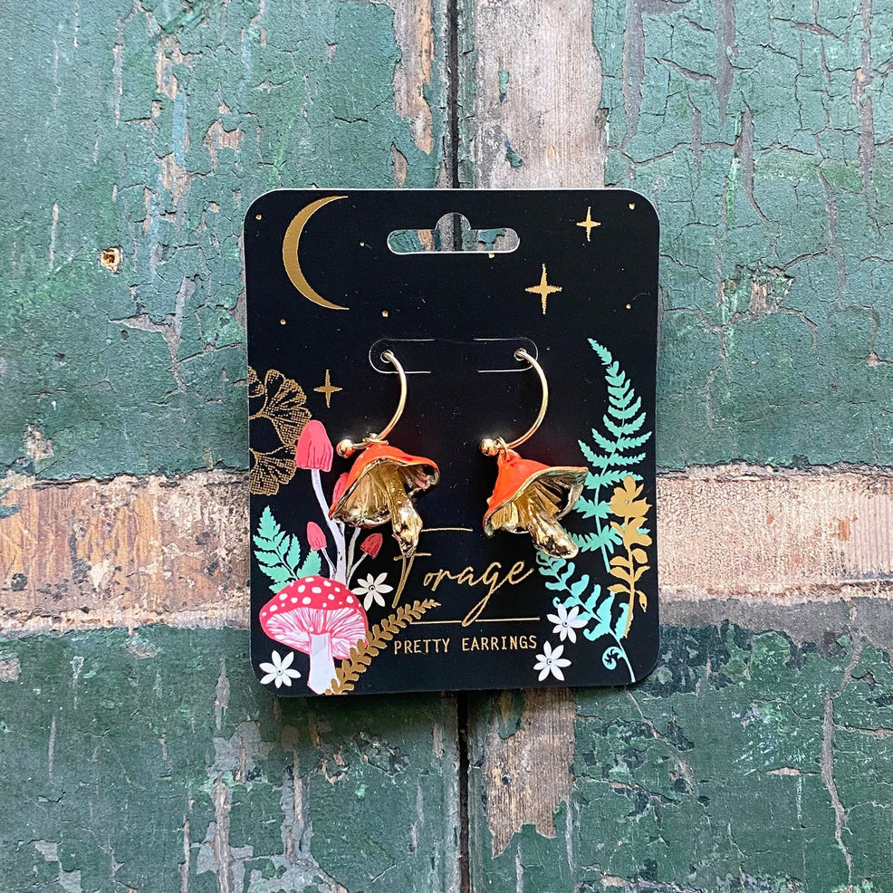 Forage Red Earrings displayed on illustrated black backing card with gold foil, showing whimsical mushroom design with gold hoops and red enamel caps.