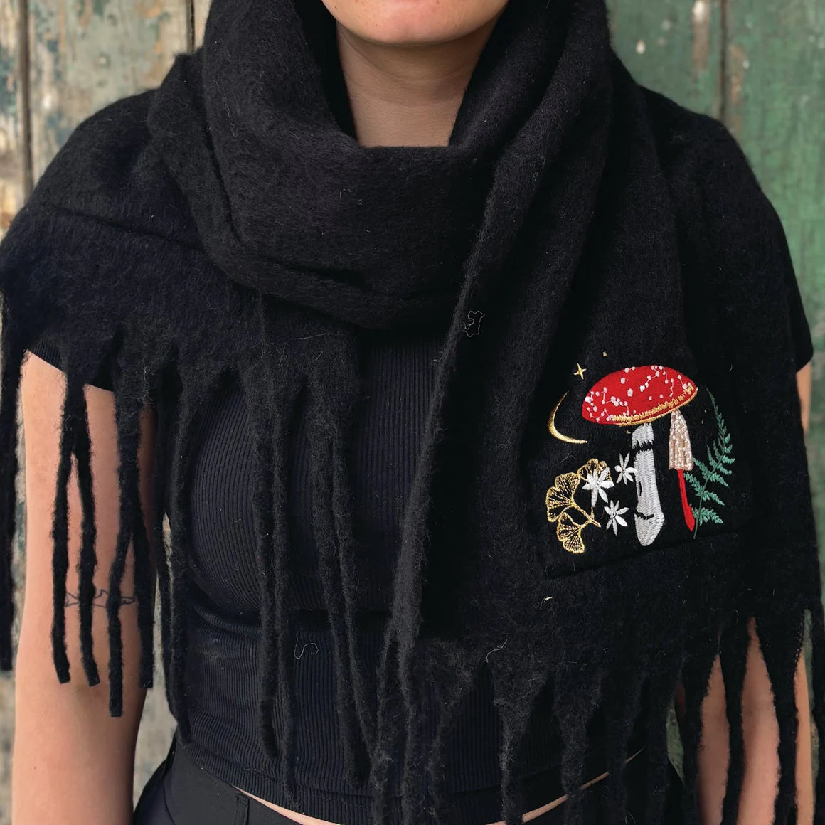 Front view of person wearing the black Forage Scarf with embroidered red toadstool and forest design, wrapped around the neck with fringe detail showing.