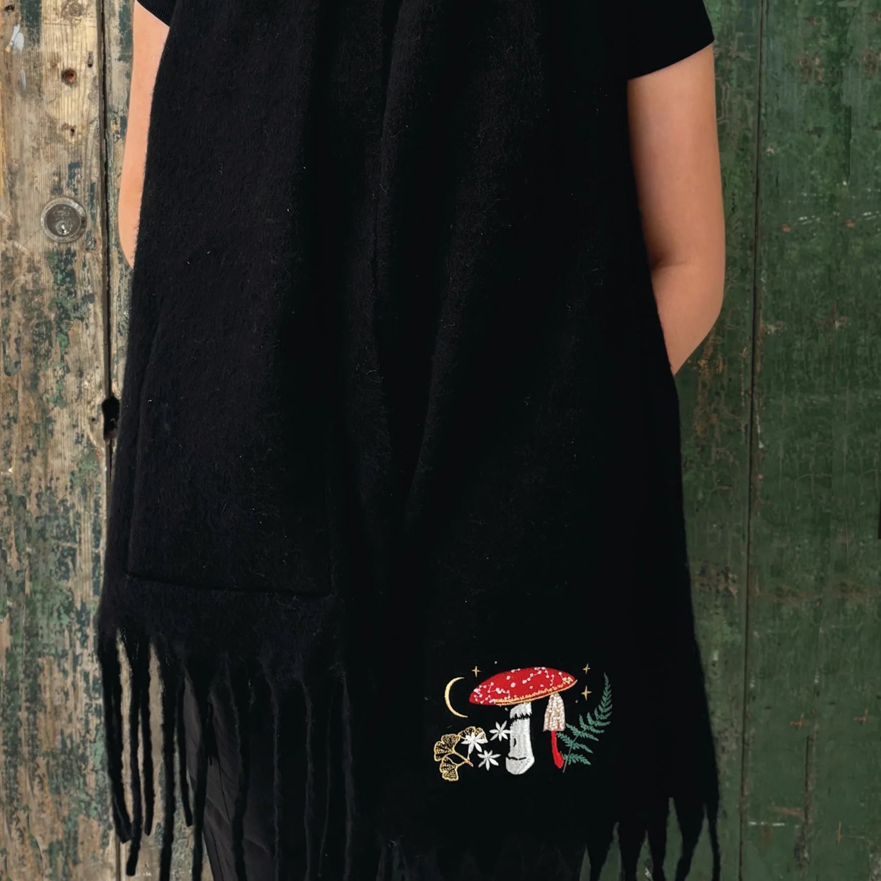 Back view of black embroidered scarf with hands in pockets and detailed woodland motif featuring mushroom, stars, and leaves.